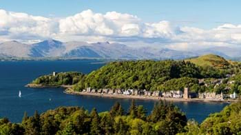 A distant view of Dunollie Wood over the hills behind Oban esplanade with buildings along the shoreline and mountains in the background